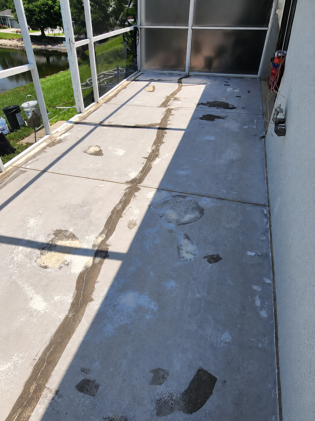 Stained and patched concrete patio floor inside a screened lanai, with grass and water visible through the screen enclosure